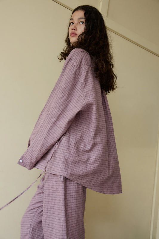 A woman with curly hair looks over her shoulder while wearing the loose-fitting String Linen Set - Musk Check by Deiji Studios, standing indoors against a cream-colored wall.