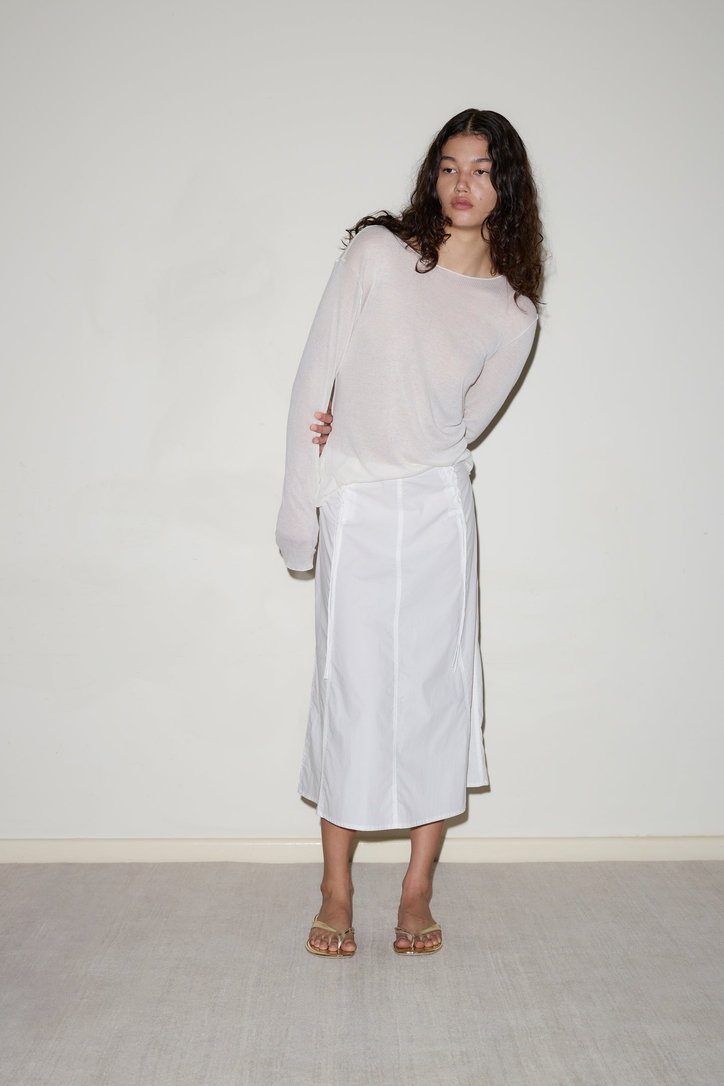 A woman with long curly hair wears the Knit Long Sleeve - Off White, a white midi skirt, and sandals while standing against a plain light-colored wall and carpeted floor.