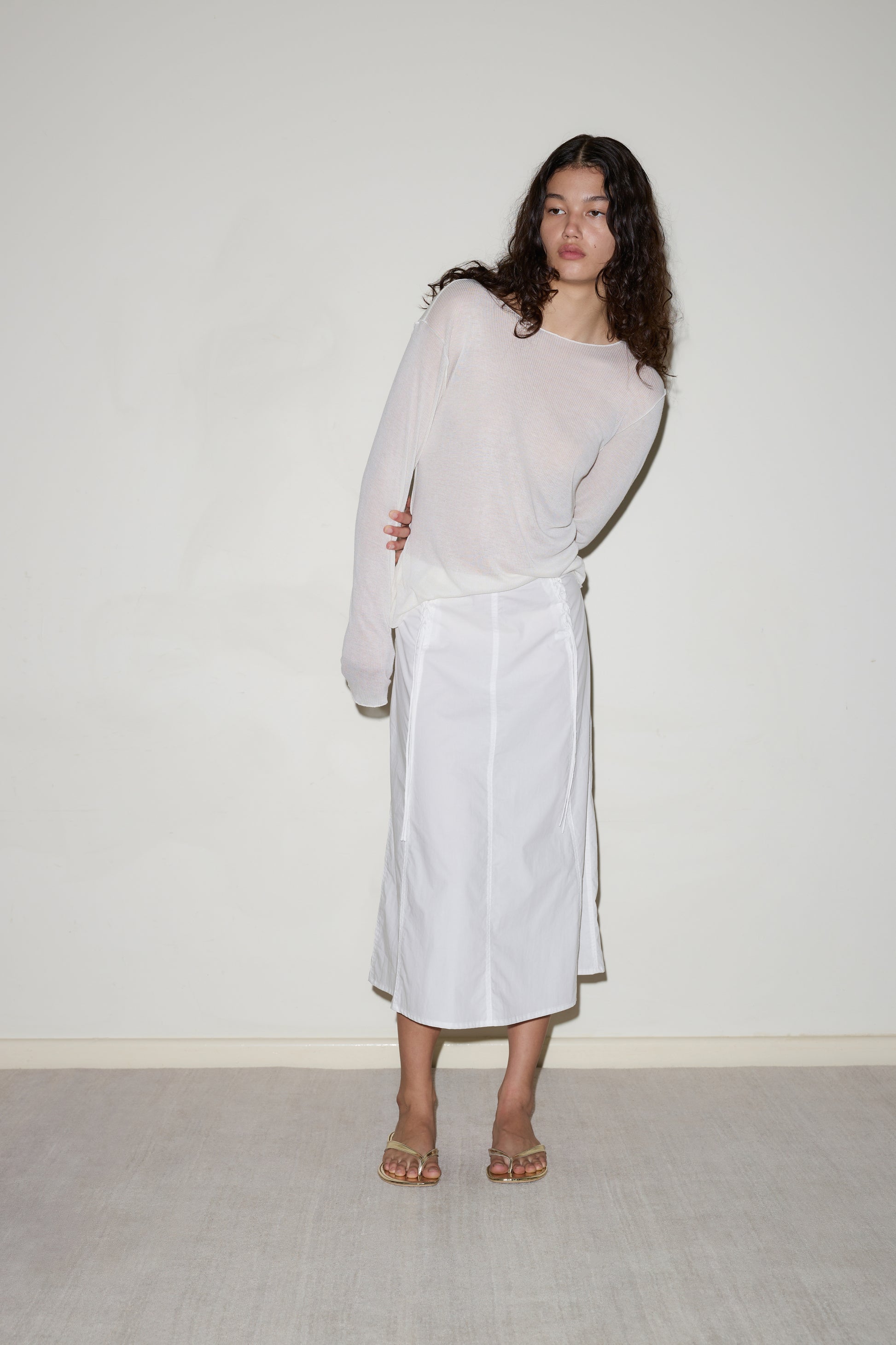 A woman with long curly hair wears the Knit Long Sleeve - Off White, a white midi skirt, and sandals while standing against a plain light-colored wall and carpeted floor.