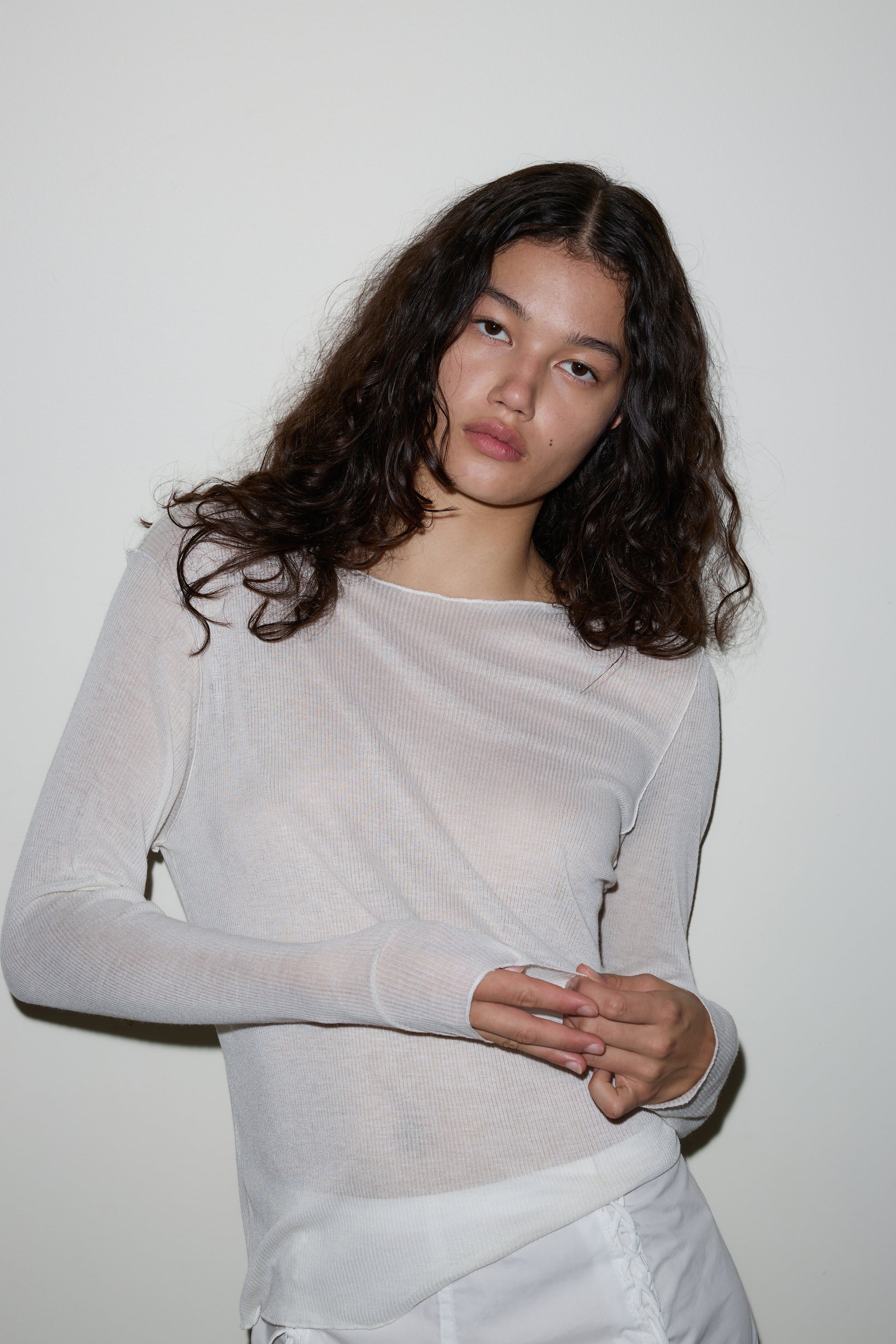 A woman with long, wavy dark hair wears a sheer white Knit Long Sleeve and white pants, standing against a plain white wall and looking directly at the camera with a neutral expression.