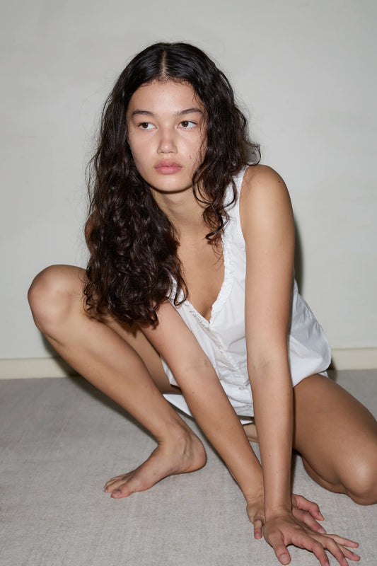A young woman with long curly hair wears a sleeveless white PJ Short Set as she crouches on the floor, gazing to the side with a neutral expression against a simple, light-colored background.