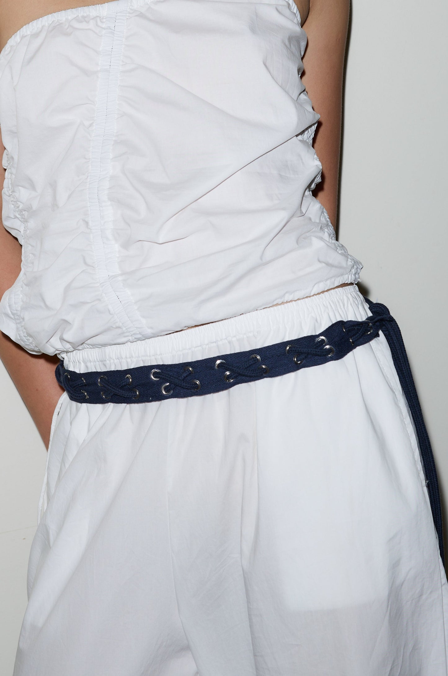 A person stands against a white background wearing a white sleeveless ruched top, loose white pants, and the Eyelet Belt - Navy.