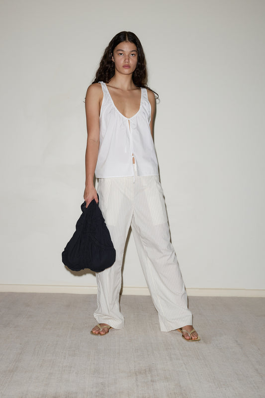 A woman stands indoors against a plain background, wearing the cream Twin Button Pant with a loose white sleeveless top, sandals, and a large black bag. She has long curly hair.