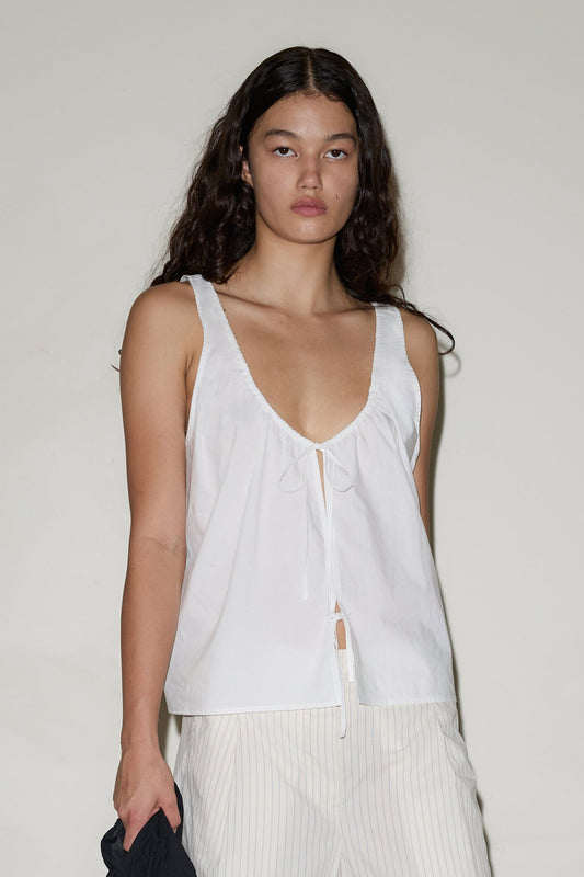 A person with long dark hair wears the Cotton Bind Top, a sleeveless white top with a low neckline and tie detail, paired with light-striped pants, standing against a plain light background.