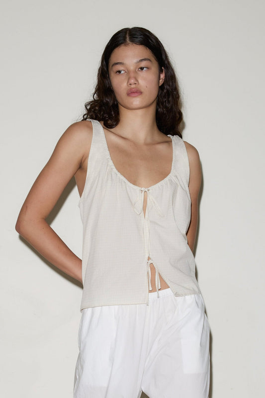 A woman with long, dark wavy hair models the Cotton Bind Top - Bone Check by Deiji Studios— a sleeveless, loose organic cotton poplin top with a front tie—paired with white pants against an off-white background.