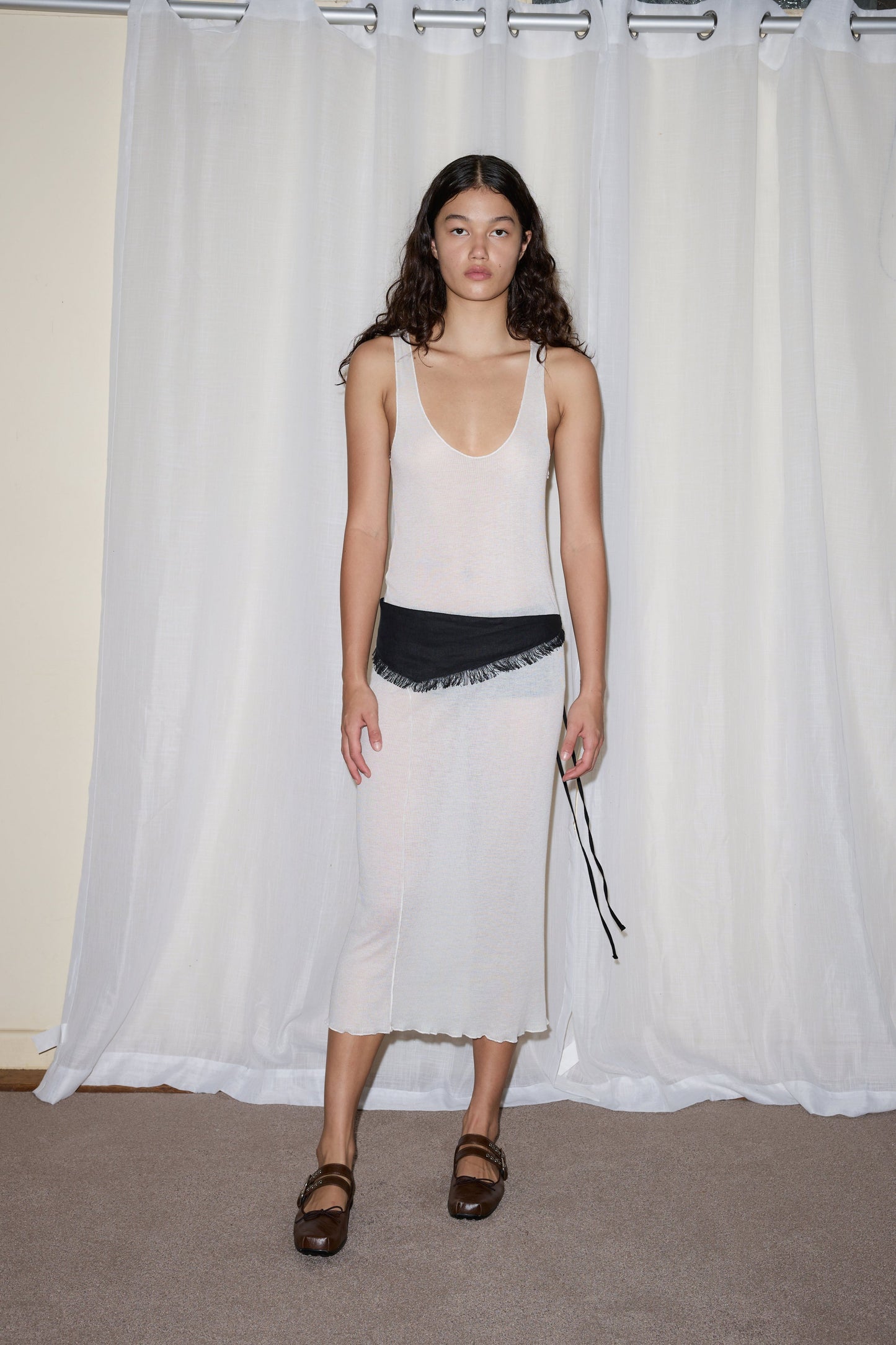 A woman with long curly hair wears the Knit Singlet Dress - Off White, paired with brown sandals and a black fringed belt bag. She stands on a carpeted floor in front of white curtains.