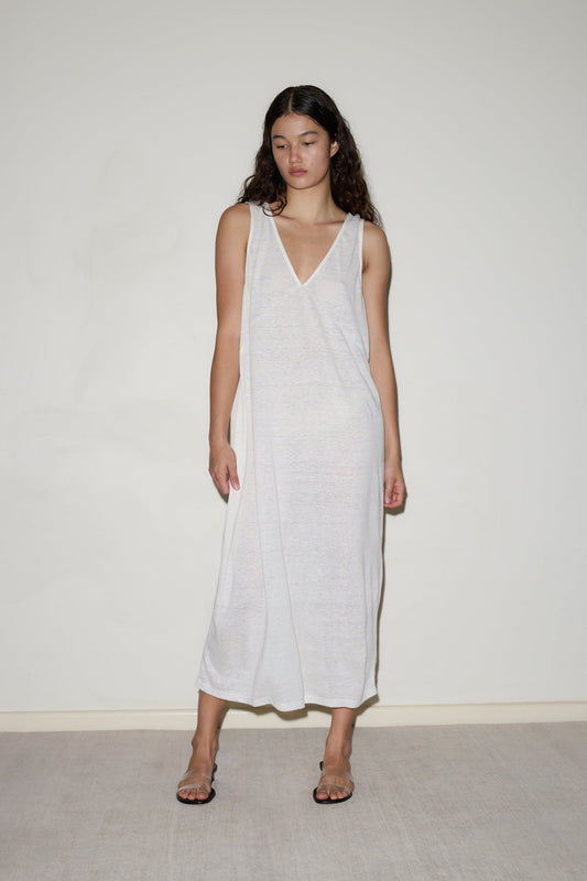 A woman with long dark hair models the Soft Tank Dress, a sleeveless, loose-fitting, semi-sheer white piece, paired with clear heels against a plain light-colored wall.