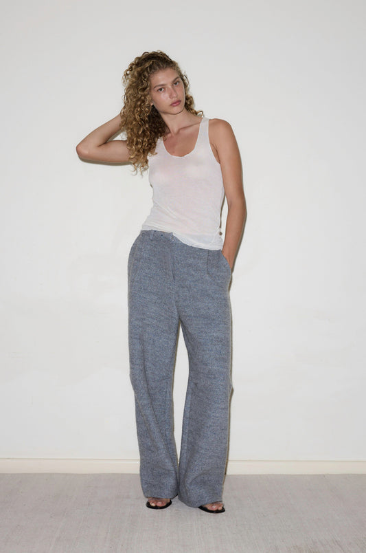 A woman with curly hair wears Boiled Wool Pants in gray and a white tank top, standing against a plain white wall. One hand is in her pocket while the other touches her hair as she looks slightly to the side.
