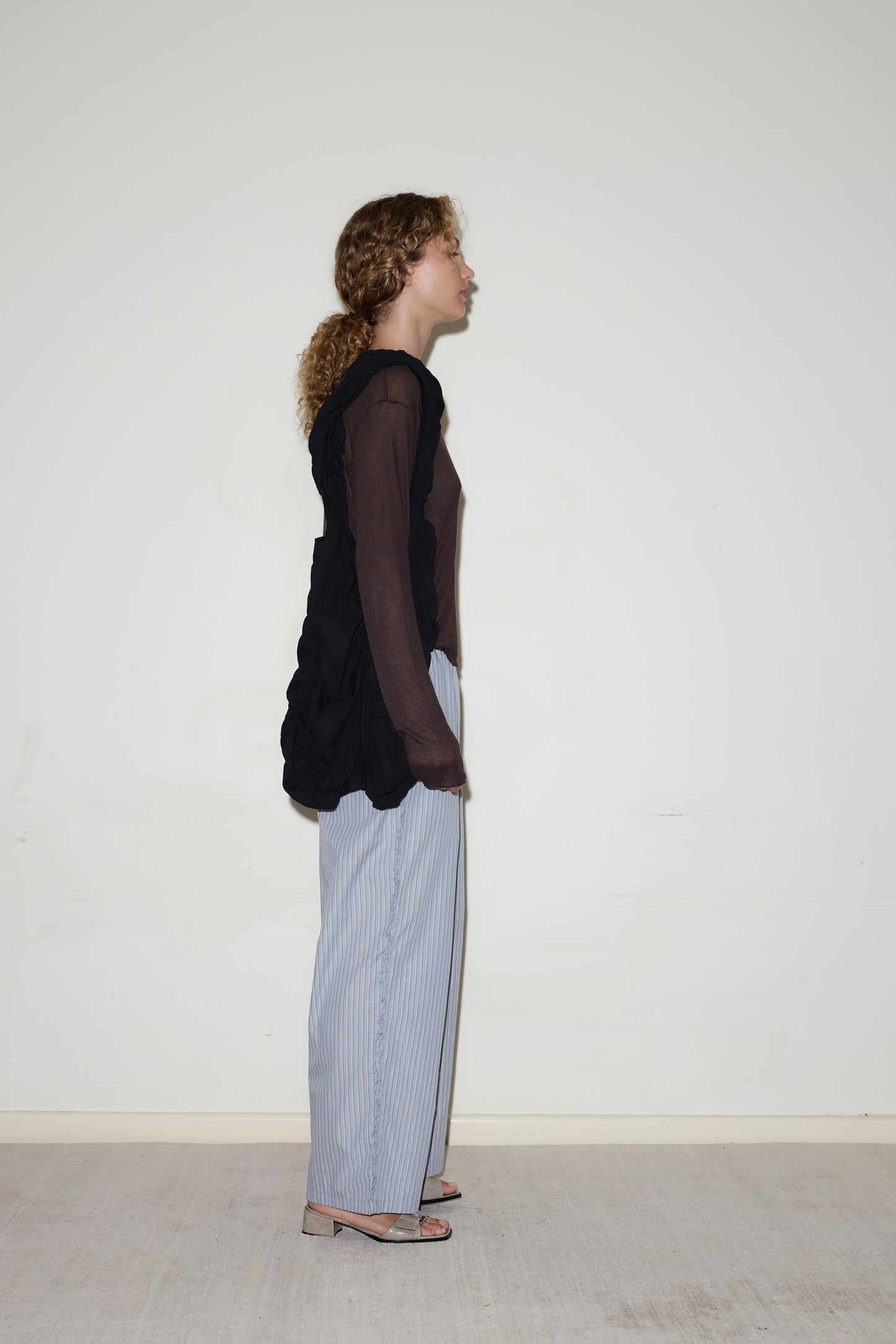 A person with curly hair in a ponytail stands in profile against a plain white wall, wearing a Deiji Studios Knit Long Sleeve in Mahogany, light blue striped pants, and open-toe heels.