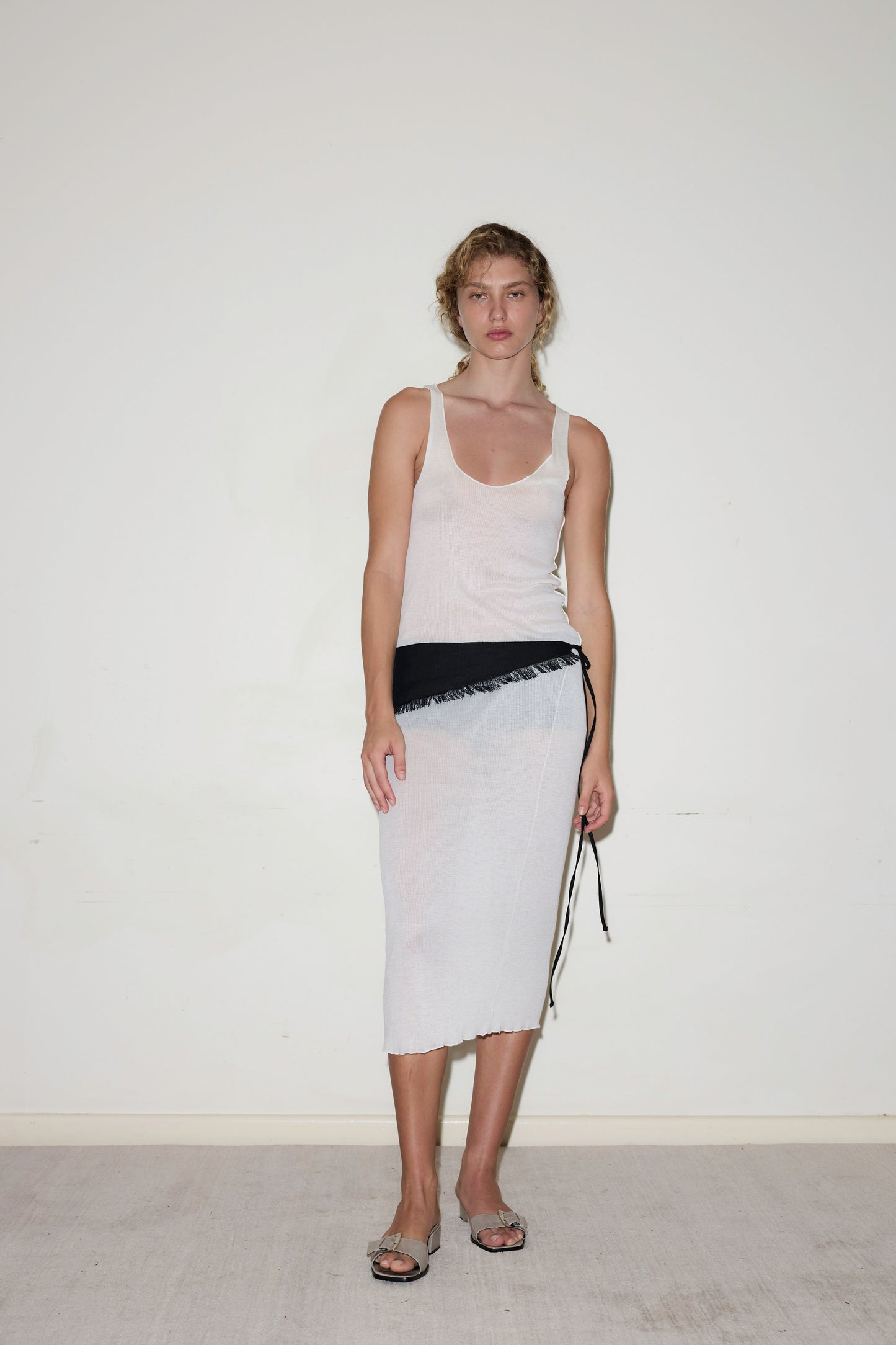 A woman with curly hair tied back and a neutral expression wears the Knit Singlet Dress in Off White, styled with a black belt and silver sandals, standing against a plain white wall.
