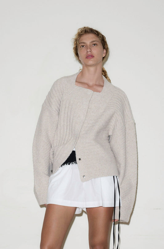 A person models the OS Square Cardigan, a loose beige knit sweater, paired with white shorts against a plain white background while looking into the camera with a relaxed expression.