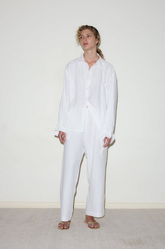 A model wears the String Linen Set—an oversized button-up shirt with tie detail and wide-leg trousers—in all white, styled with gold flat sandals against a cream background.