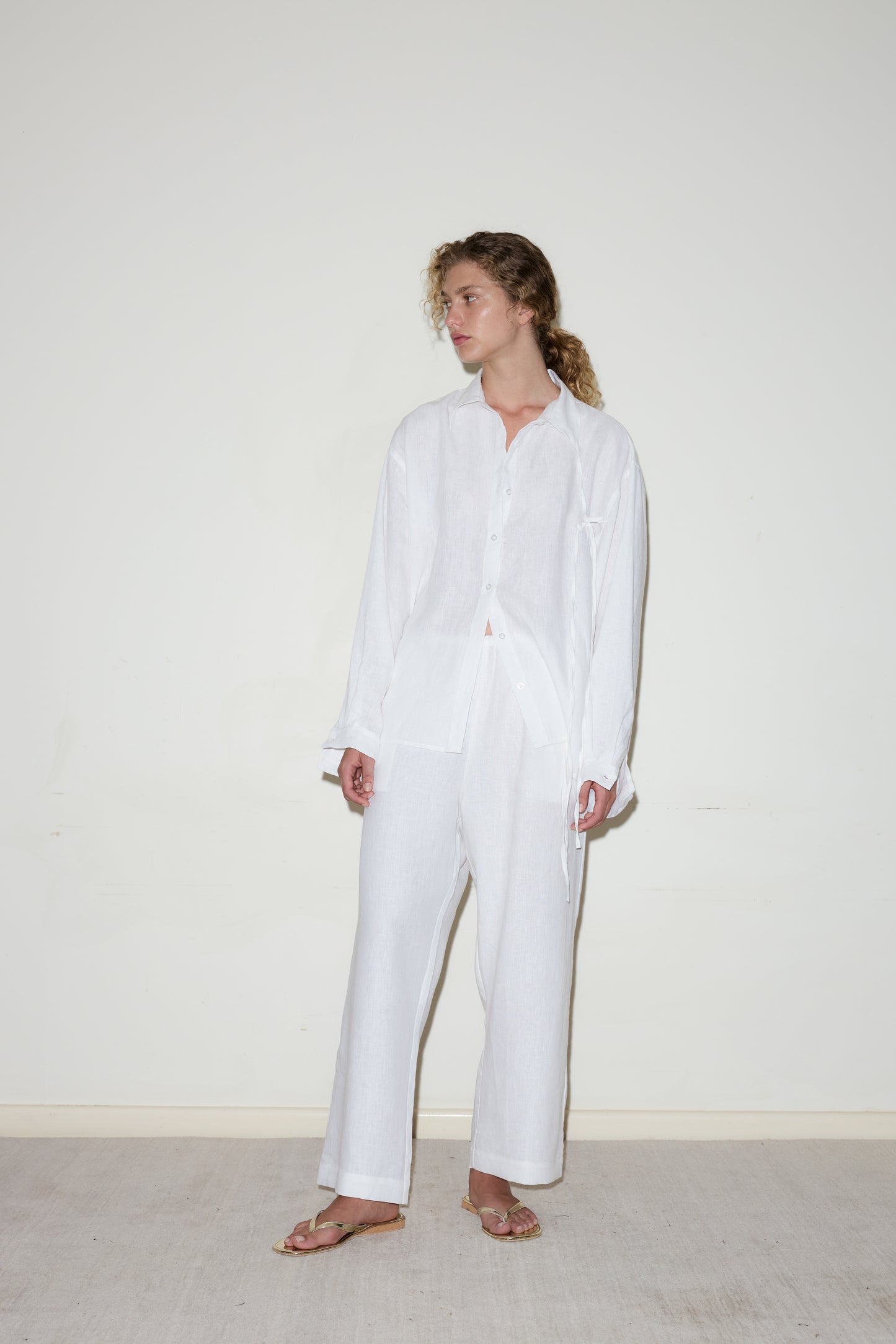 Wearing the String Linen Set - White, a person with curly hair stands barefoot against a plain off-white wall, gazing to the side.