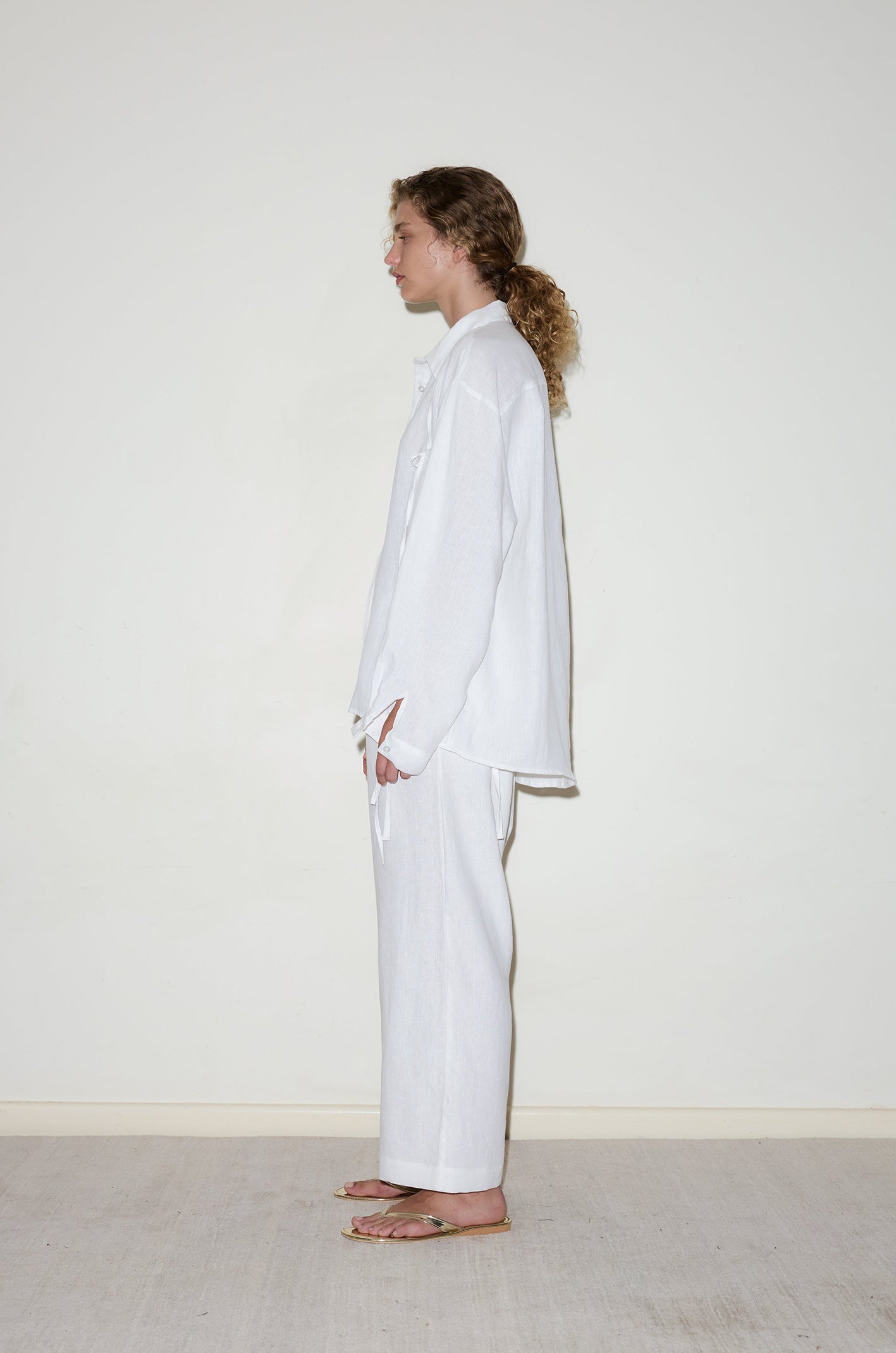 A person with curly hair tied back stands in profile against a plain off-white wall, wearing the String Linen Set - White and brown sandals.