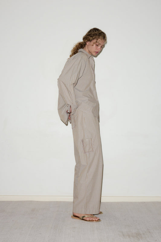 A person with curly hair stands sideways against a white wall, wearing the Cotton Pyjama Set—a loose-fitting, beige and white striped outfit with long sleeves, wide-leg pants, and brown sandals.