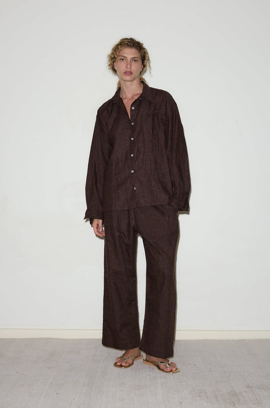 A person stands indoors against a plain white wall, wearing the String Linen Set—a loose-fitting dark brown button-up shirt and matching wide-leg pants—paired with gold sandals for a relaxed, casual look.