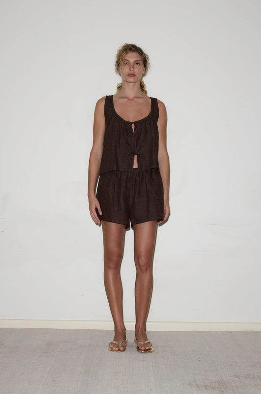 A person stands against a plain white wall, wearing the Open Linen Short Set— a sleeveless, loose-fitting dark brown top with a tie front and matching shorts. Their curly hair is pulled back, and they are wearing sandals.