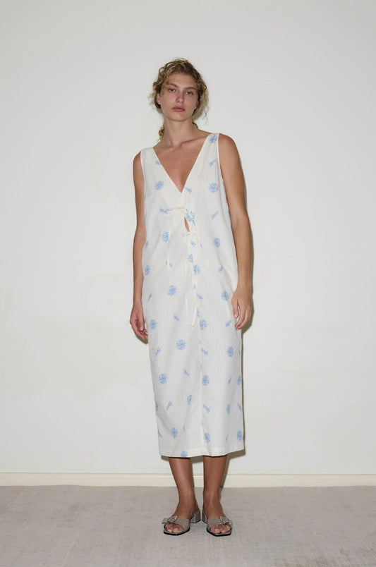 Against a plain white wall, a woman with curly hair wears the Cross Over Dress - Sea Flower, an organic cotton midi dress featuring blue floral prints, a deep V-neckline, and front tie detail. She pairs it with flat sandals.
