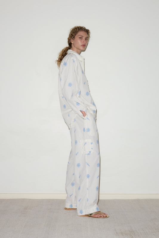 A person with curly hair stands sideways against a white wall and beige carpet, wearing the Cotton Pyjama Set— a loose white outfit featuring light blue floral patterns—paired with flip-flops.