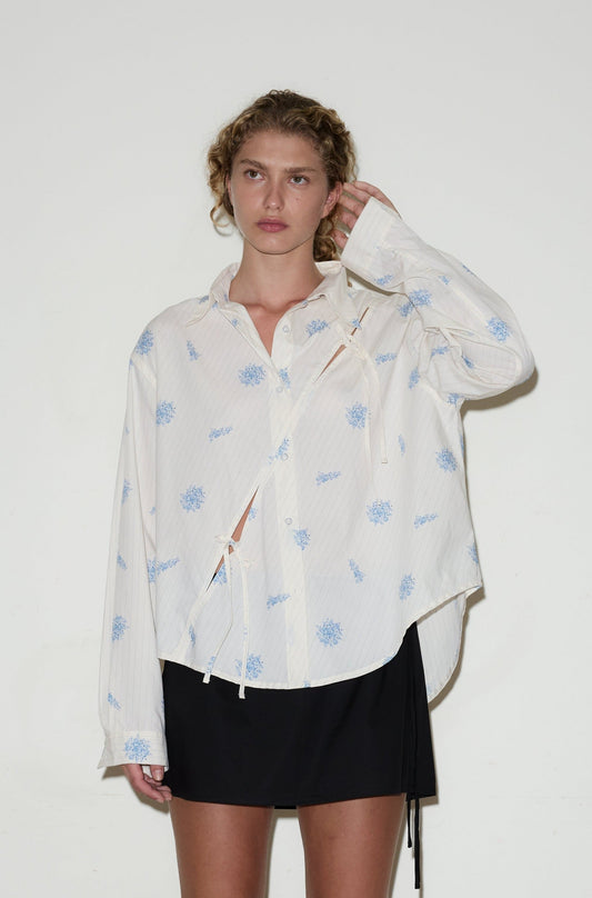A woman with curly hair wears the twin bow shirt, featuring a loose fit and light blue floral patterns, paired with a short black skirt. She stands against a plain white background, touching her hair with one hand.