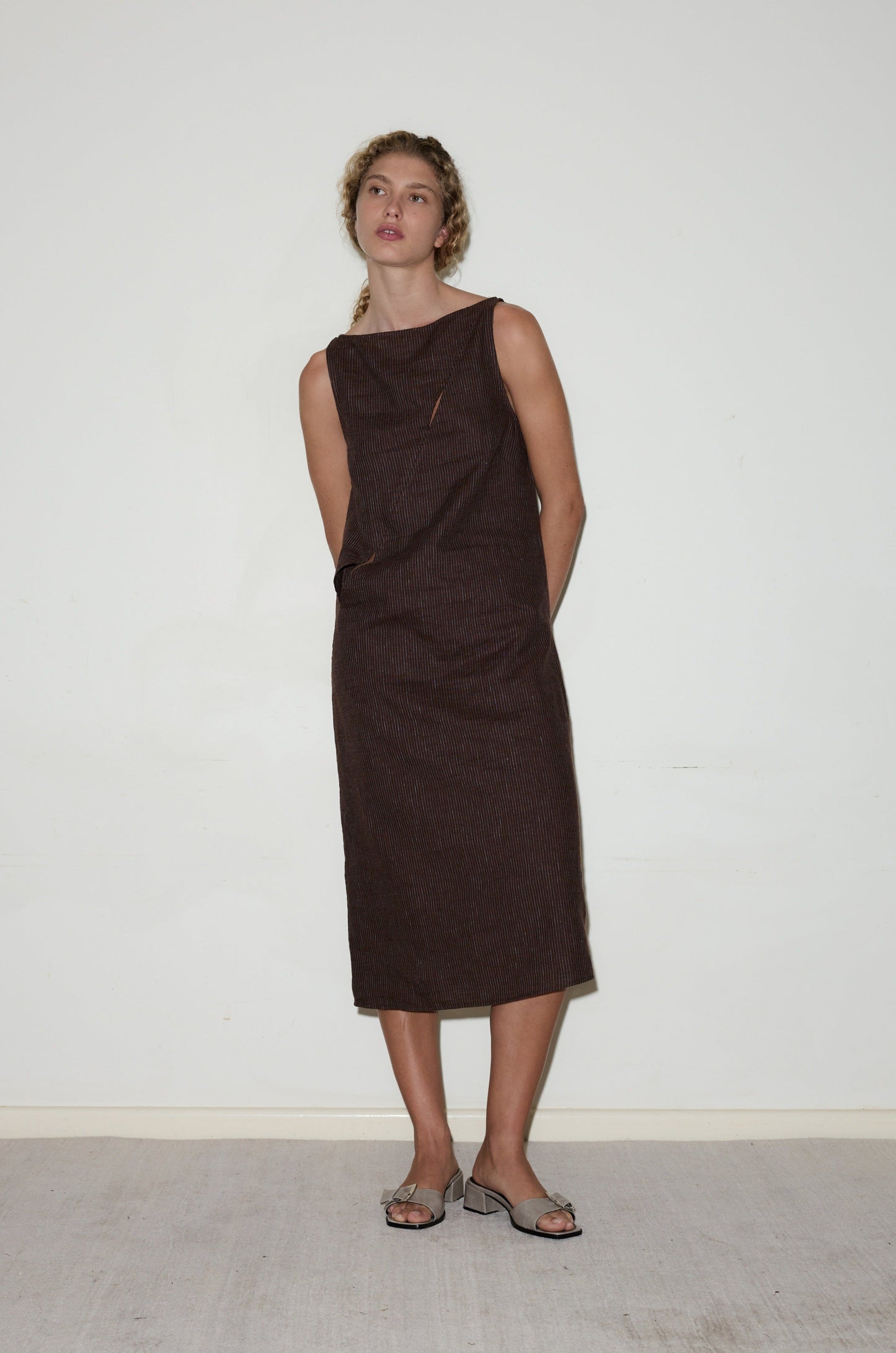 A person stands against a white wall, wearing the sleeveless, knee-length High Tie Dress in dark brown with light open-toe sandals. Their hands are behind their back and their hair is styled in loose waves.