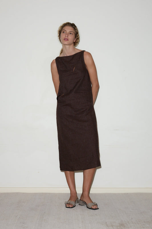 A person with curly hair, wearing the High Tie Dress—a dark brown, sleeveless, knee-length outfit—and open-toed heeled sandals stands against a plain white wall with hands behind their back.