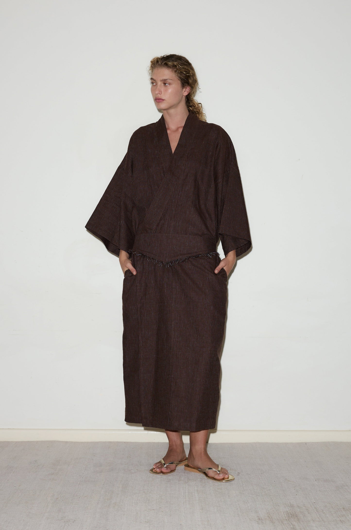 A person stands against a plain white wall, wearing Deiji Studios’ 02 Robe - Royal Stripe with wide sleeves and a long skirt, paired with brown sandals. Their hands are in their pockets as they look to the side.