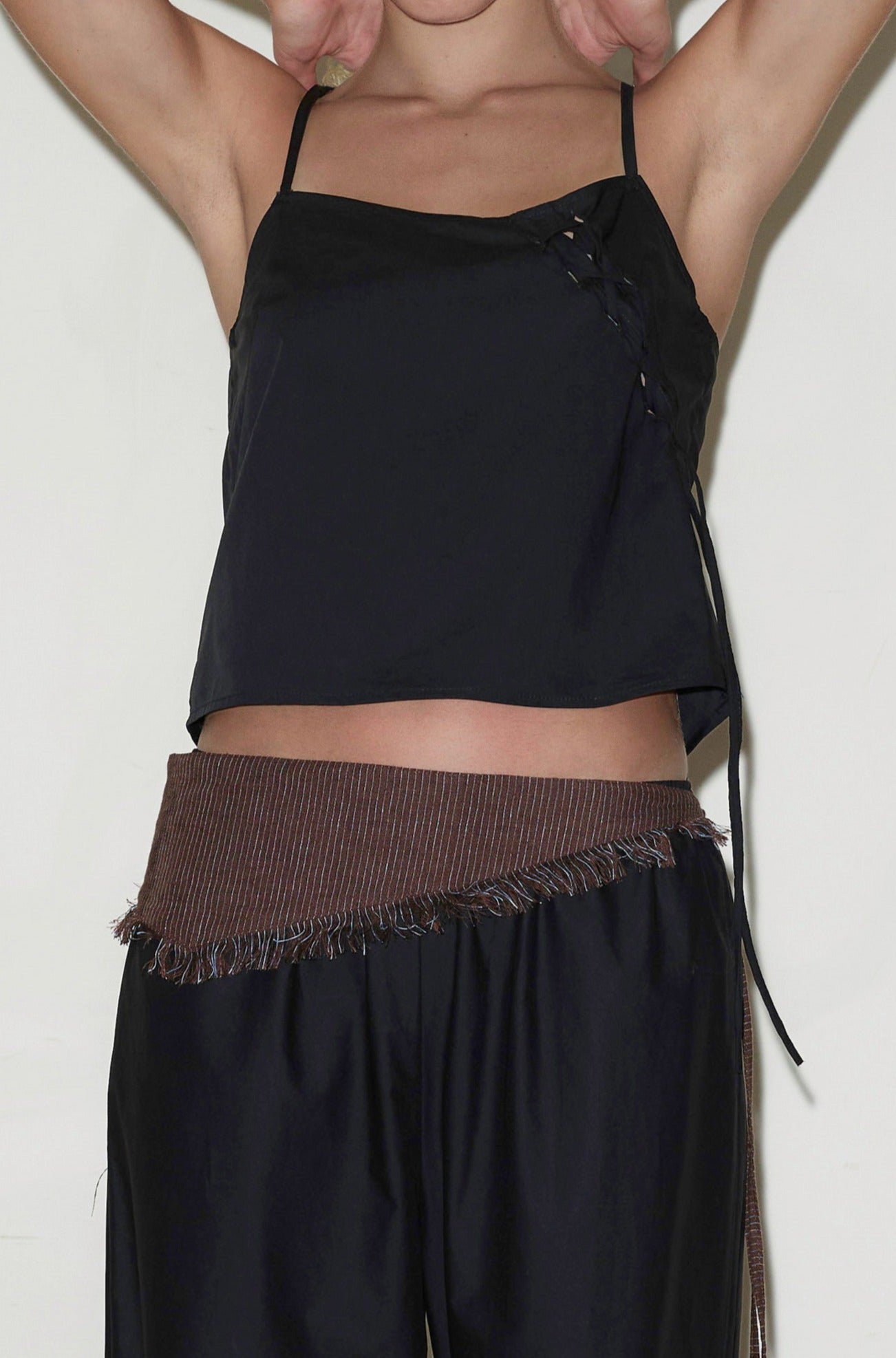 Wearing the Eyelet Top - Black with lace-up sides and black pants, paired with a brown fringed, asymmetrical belt; arms raised and face not visible.