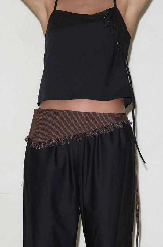 A person wears a sleeveless black top with thin straps and side lace-up detail, loose black pants, and the Linen Fray Belt—a brown belt with frayed edges—around the waist. The head and lower legs are not visible.