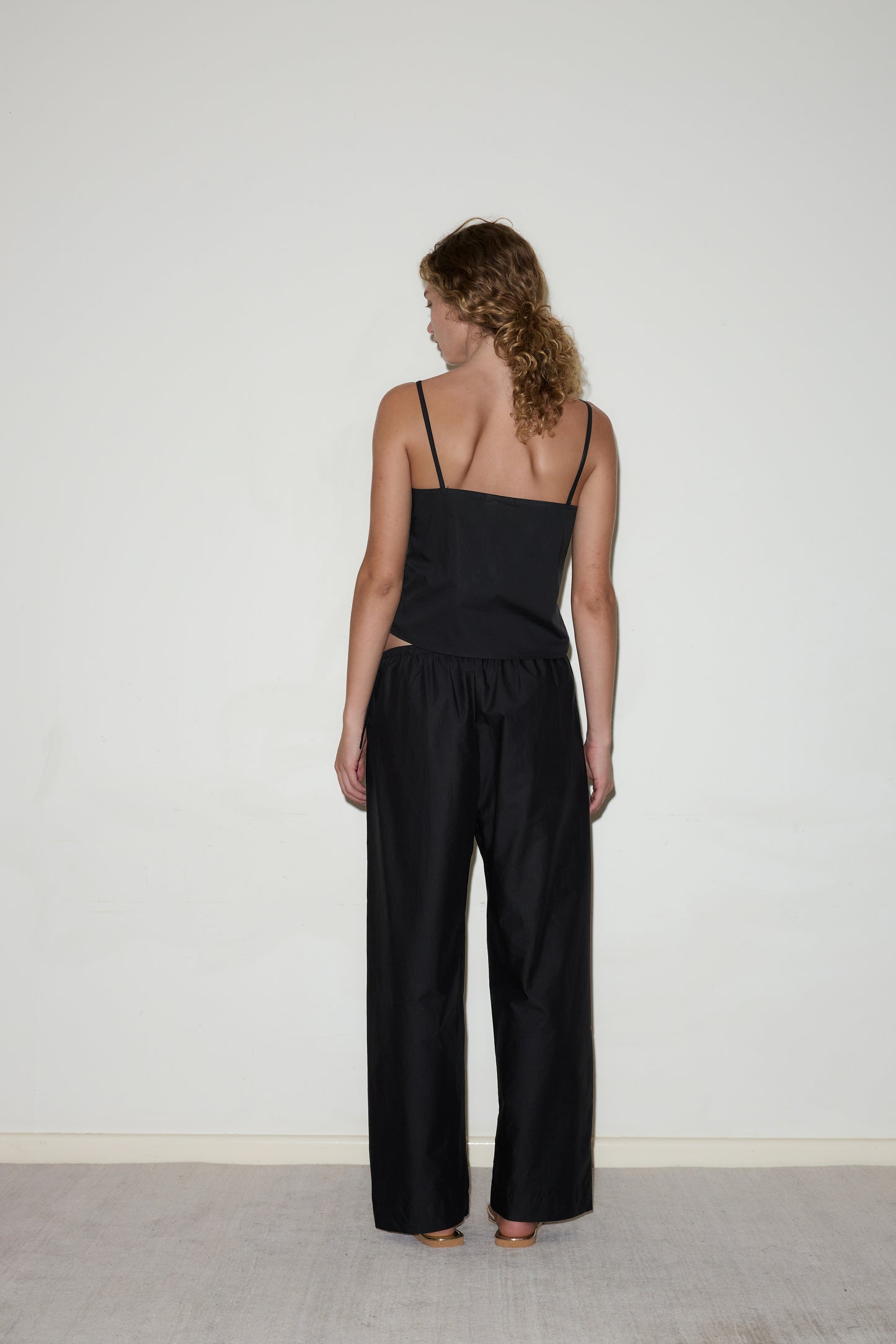 A woman with curly hair, seen from the back facing a white wall, wears the Eyelet Top - Black by Deiji Studios and loose black pants.