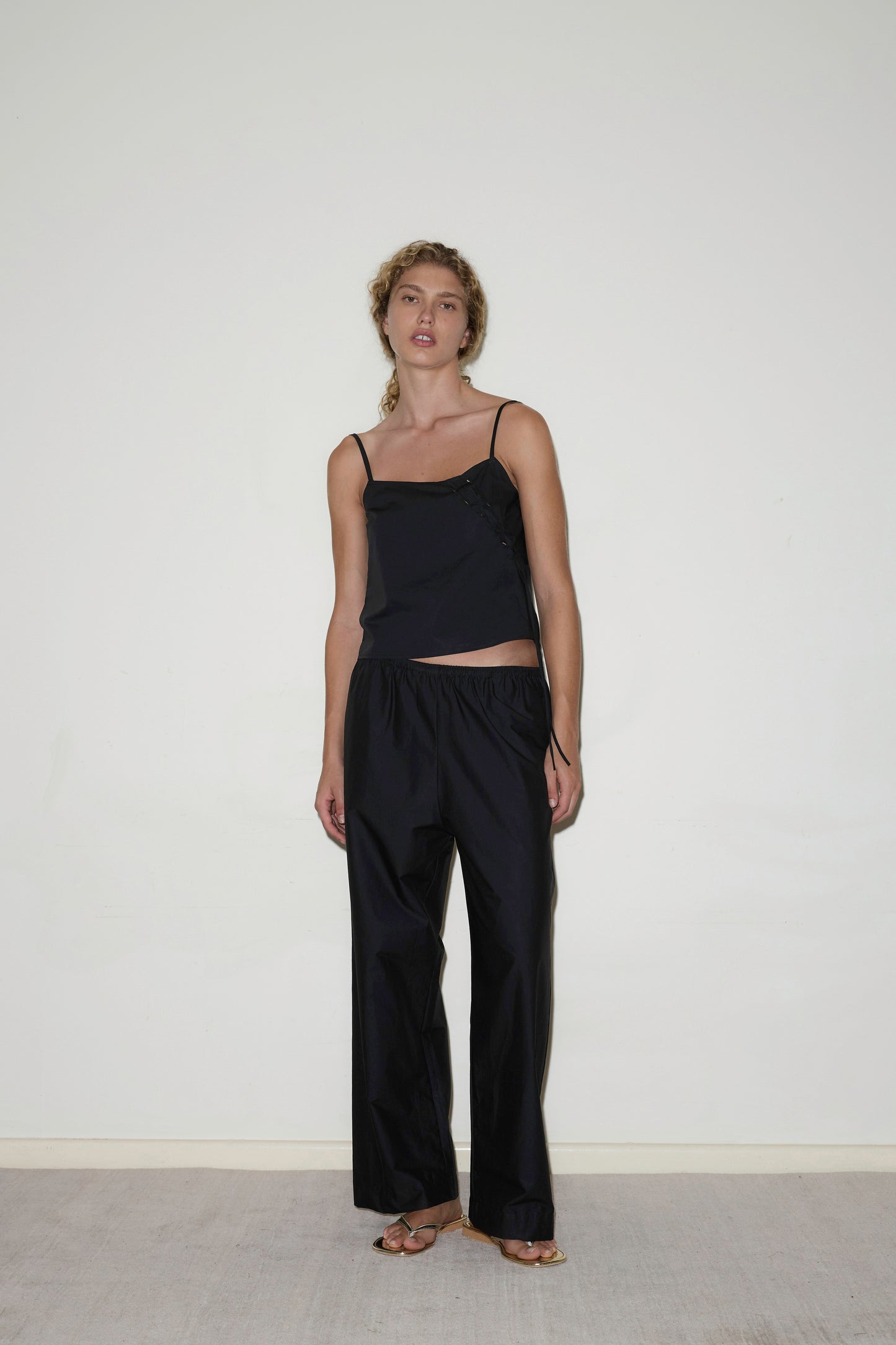 A woman stands indoors against a plain white wall, wearing the black Eyelet Top with spaghetti straps and loose black pants. She is barefoot, with a relaxed posture and neutral expression.