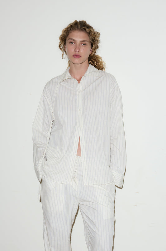 A person with curly hair wears a loose-fitting, light-colored Lapel Bow Shirt and stands against a plain white wall with hands in their pockets, looking at the camera with a neutral expression.