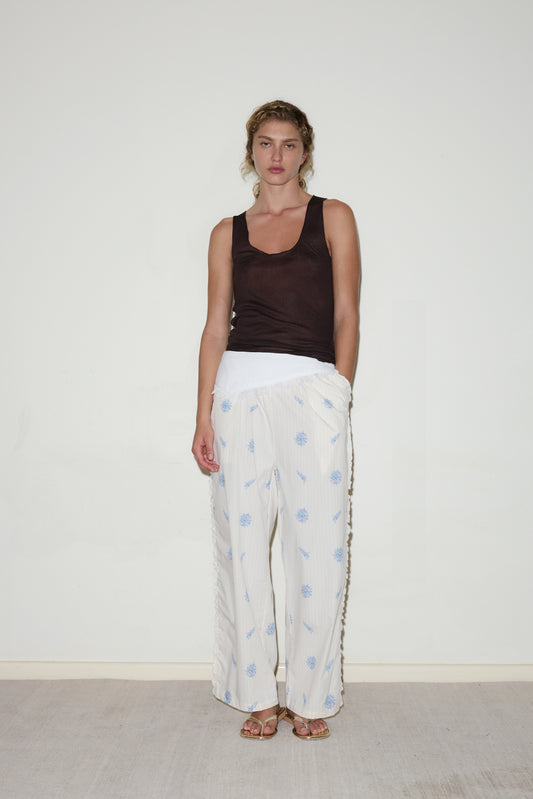 A woman stands against a white wall wearing a sleeveless dark brown top, the Ruffle Pant - Sea Flower with light blue flower patterns by Deiji Studios, and sandals. One hand is in her pocket and she has a neutral expression.