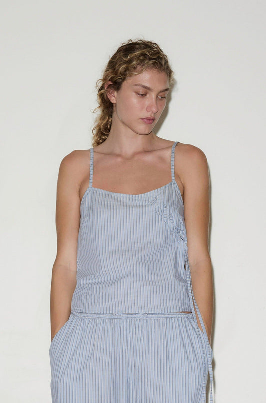 A woman with wavy blonde hair wears an Eyelet Top and matching pants, both light blue and white with vertical stripes, as she stands against a plain white background looking down with her hands in her pockets.
