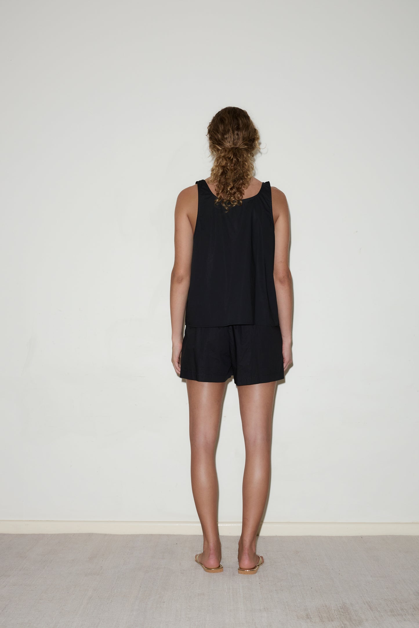 A woman with curly hair stands barefoot on a light-colored floor, facing a white wall. She wears Deiji Studios’ Cotton Bind Top - Black and black shorts, her arms relaxed at her sides.