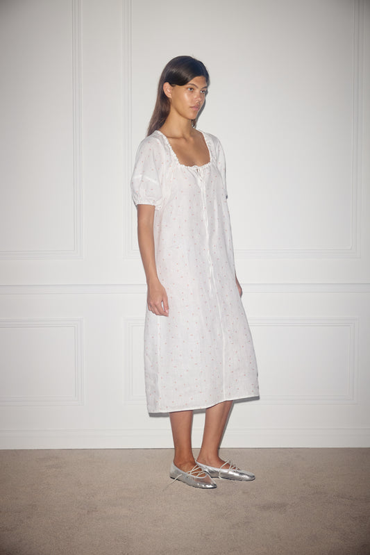 A woman stands indoors against a white paneled wall, wearing the Curved Seam Midi Dress - Corsage Print by Deiji Studios with short sleeves and a loose fit, paired with silver lace-up ballet flats.