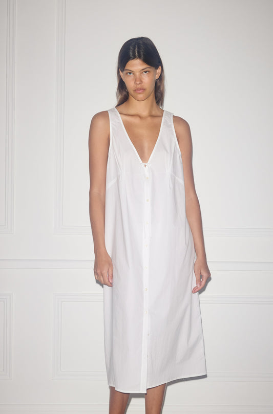 A woman with straight dark hair and a neutral expression stands against a white paneled wall, wearing the Tuck Tie Dress - White, a sleeveless, knee-length dress featuring a deep V-neckline.
