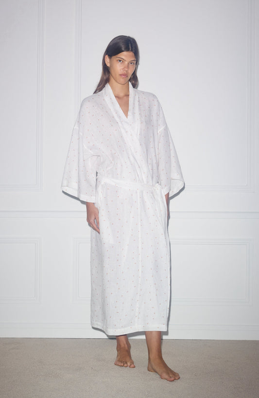 A person stands indoors against a white wall, wearing the 02 Robe—long, white, with small colored dots, wide sleeves, and a belted waist. Barefoot and neutral-faced.