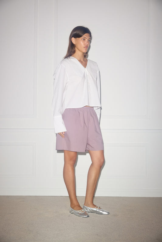 A woman stands indoors by a white paneled wall, wearing a loose white long-sleeve shirt, silver flats, and light purple mid shorts. She gazes slightly to the side with a neutral expression.