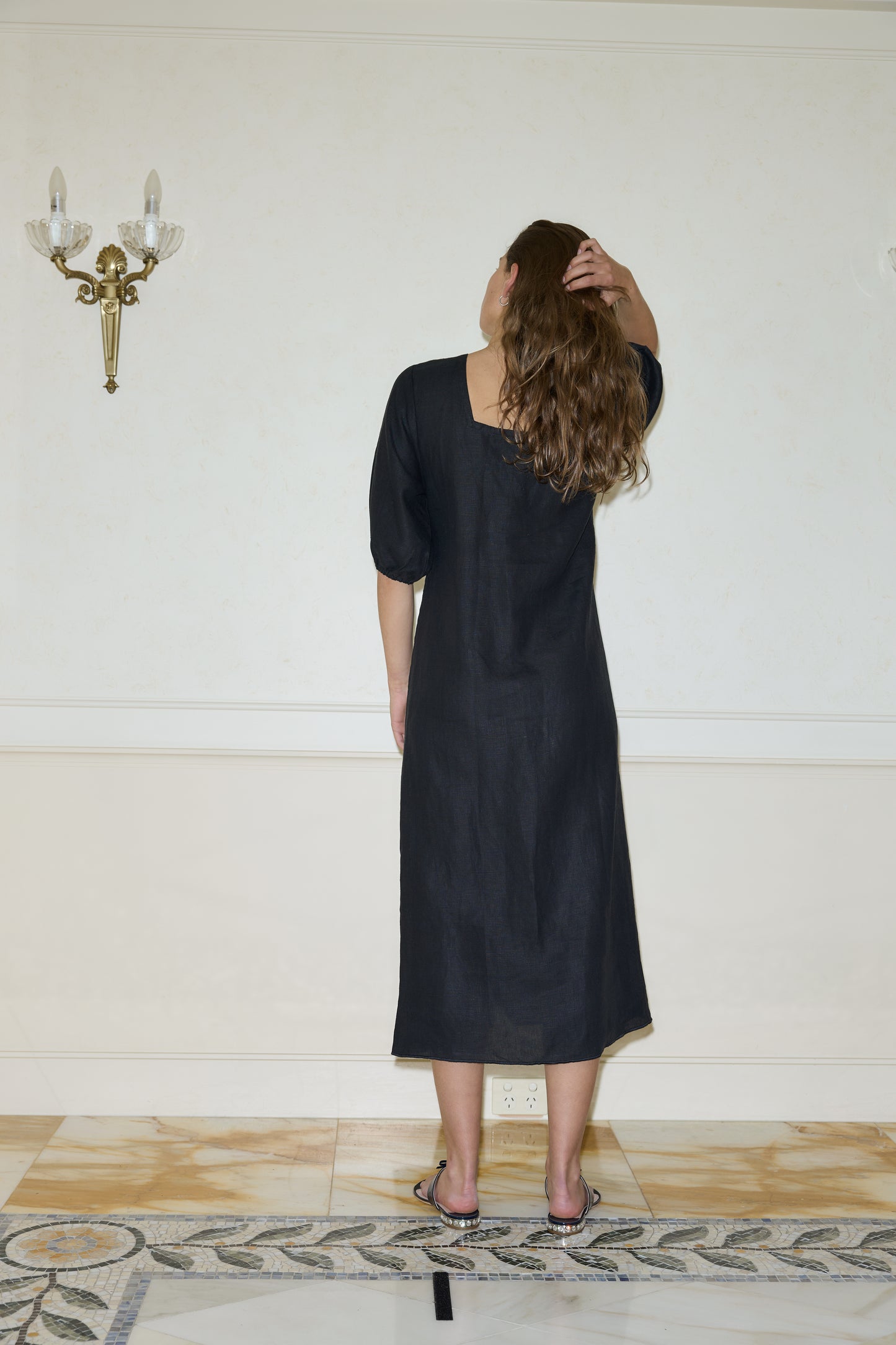 A person with long, wavy brown hair is indoors facing a white wall, wearing the Squared Dress - Black—a loose-fitting dress made of organic linen—with one hand touching the back of their head.