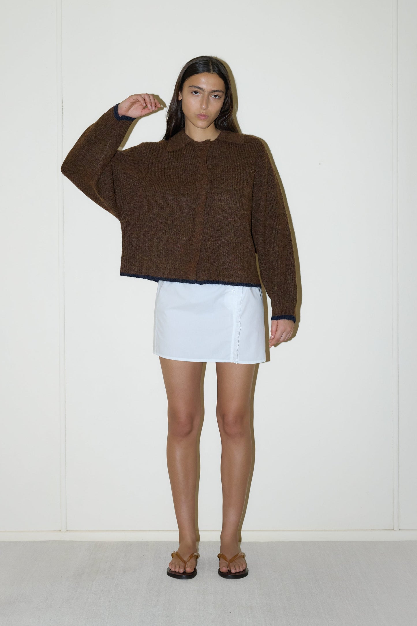dark haired female model wears a brown elevated in a luxurious softly weighted and subtly lustrous sustainable knit blend featuring invisible press stud closure front, a relaxed collar and dropped shoulder, a gently curved arm for soft shaping and oversized fit for layering, paired with a white simple mini skirt designed to sit at low waist features adjustable tie and ruffle edge on side with button detail
