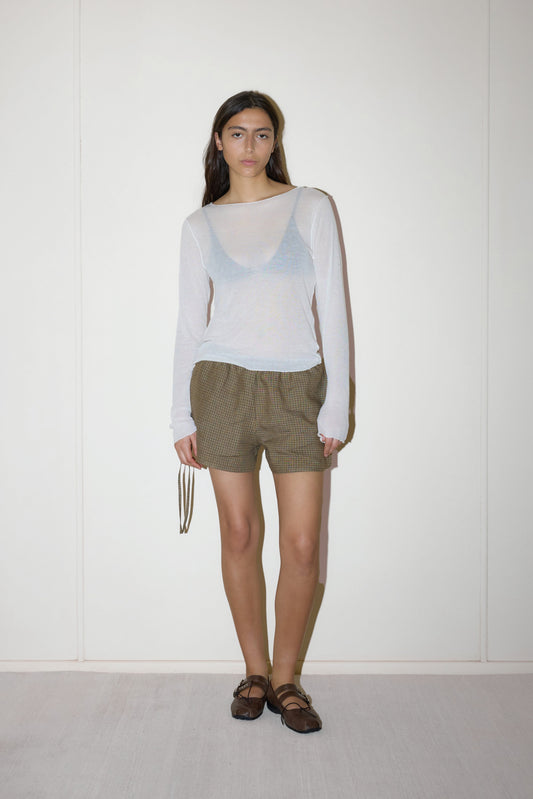 A woman stands against a white wall wearing a sheer long-sleeve top over a tank, Side Tie Short in brown plaid, and brown shoes. She holds a thin strap in one hand and looks at the camera with a neutral expression.