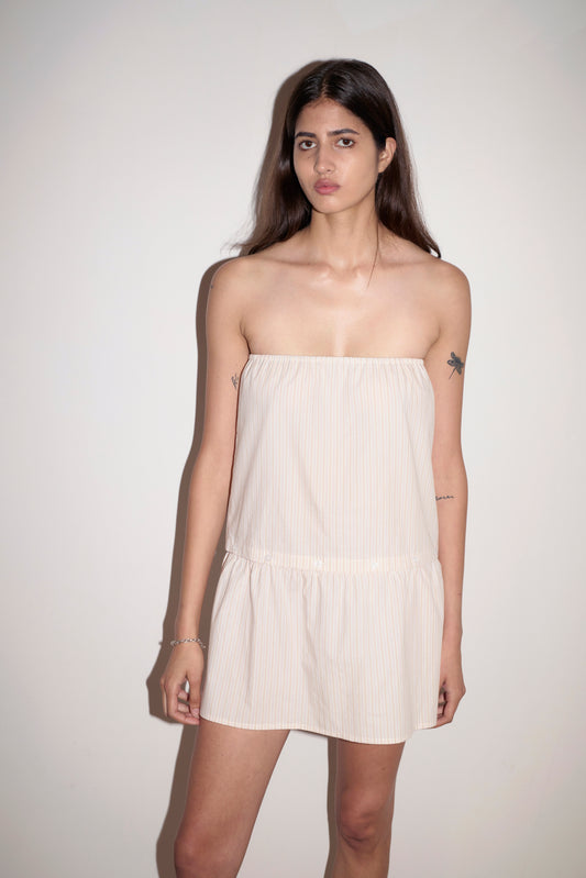 A woman with long dark hair and visible arm tattoos stands against a white wall, wearing the Button Dress—a strapless, light-toned mini dress featuring subtle vertical stripes. She has a neutral expression.