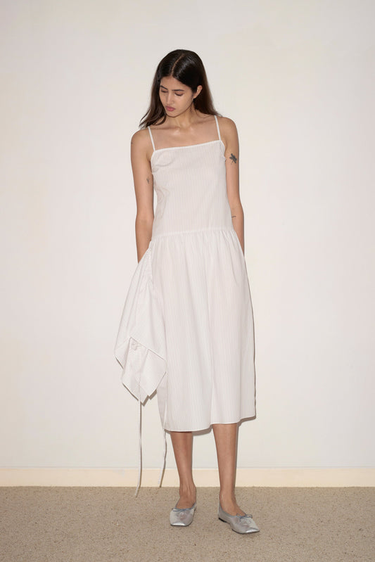 A woman with long brown hair wears the Peplum Dress—a sleeveless, white midi dress with thin straps and an asymmetrical peplum detail—paired with silver flats, standing against a plain wall and looking down.