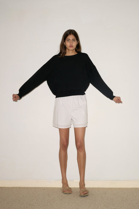 A woman stands against a plain white wall with her arms slightly outstretched, wearing the 0 Pullover - Black, white shorts, and sandals.