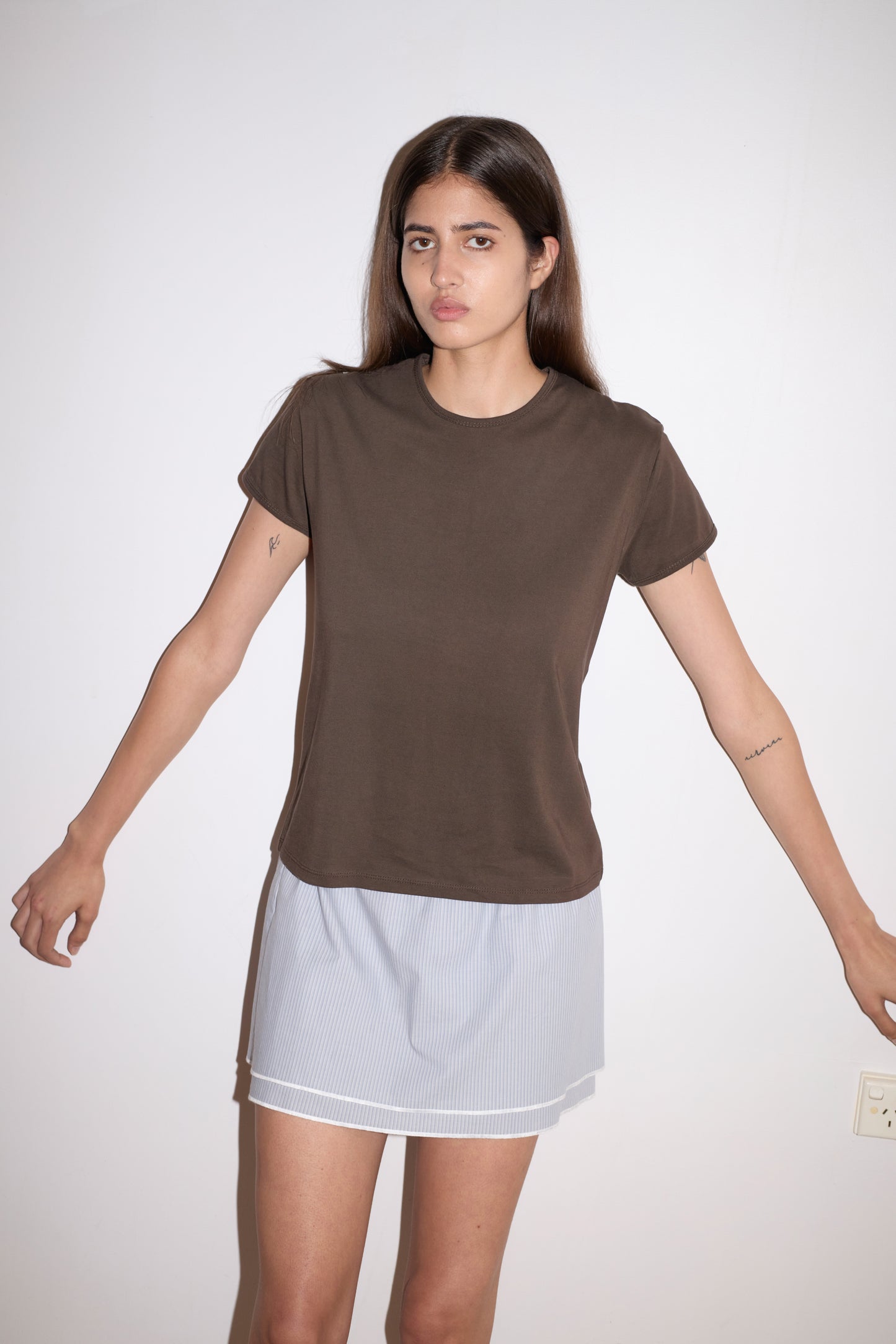 A woman with long brown hair wears the 0 Layered Skirt - Sky Stripe and a dark brown t-shirt, standing against a plain white wall with her arms slightly outstretched.