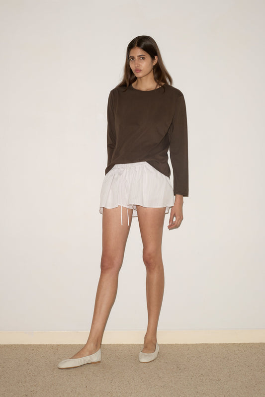 A woman stands indoors against a plain wall, wearing the Skort - Pacific Stripe by 0 and a long-sleeved brown top. She gazes slightly to the side with a neutral expression.