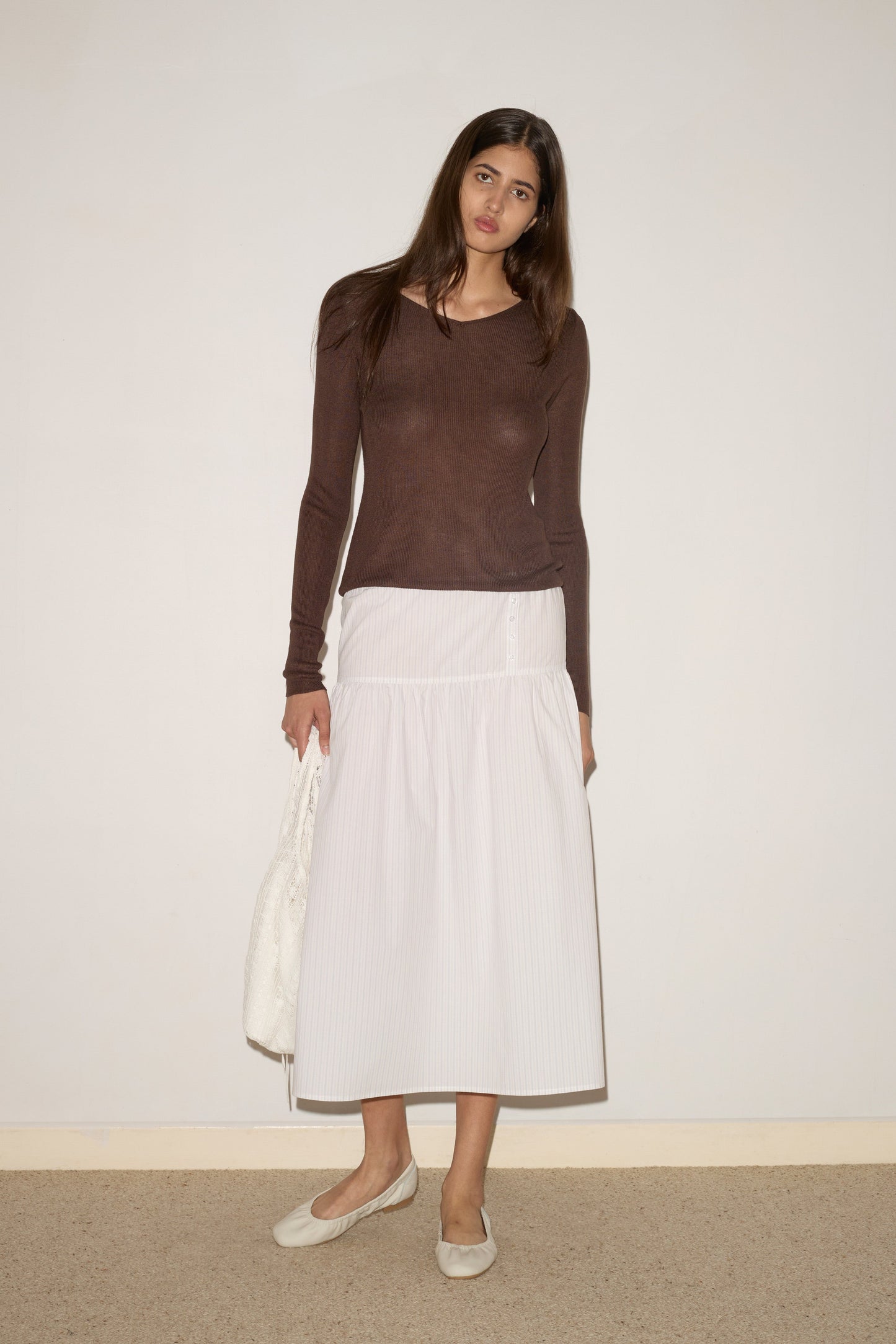 A woman with long brown hair stands by a plain wall, wearing 0’s Knitted Top in Dark Brown, a white ankle-length skirt, white flats, and holding a white textured bag.