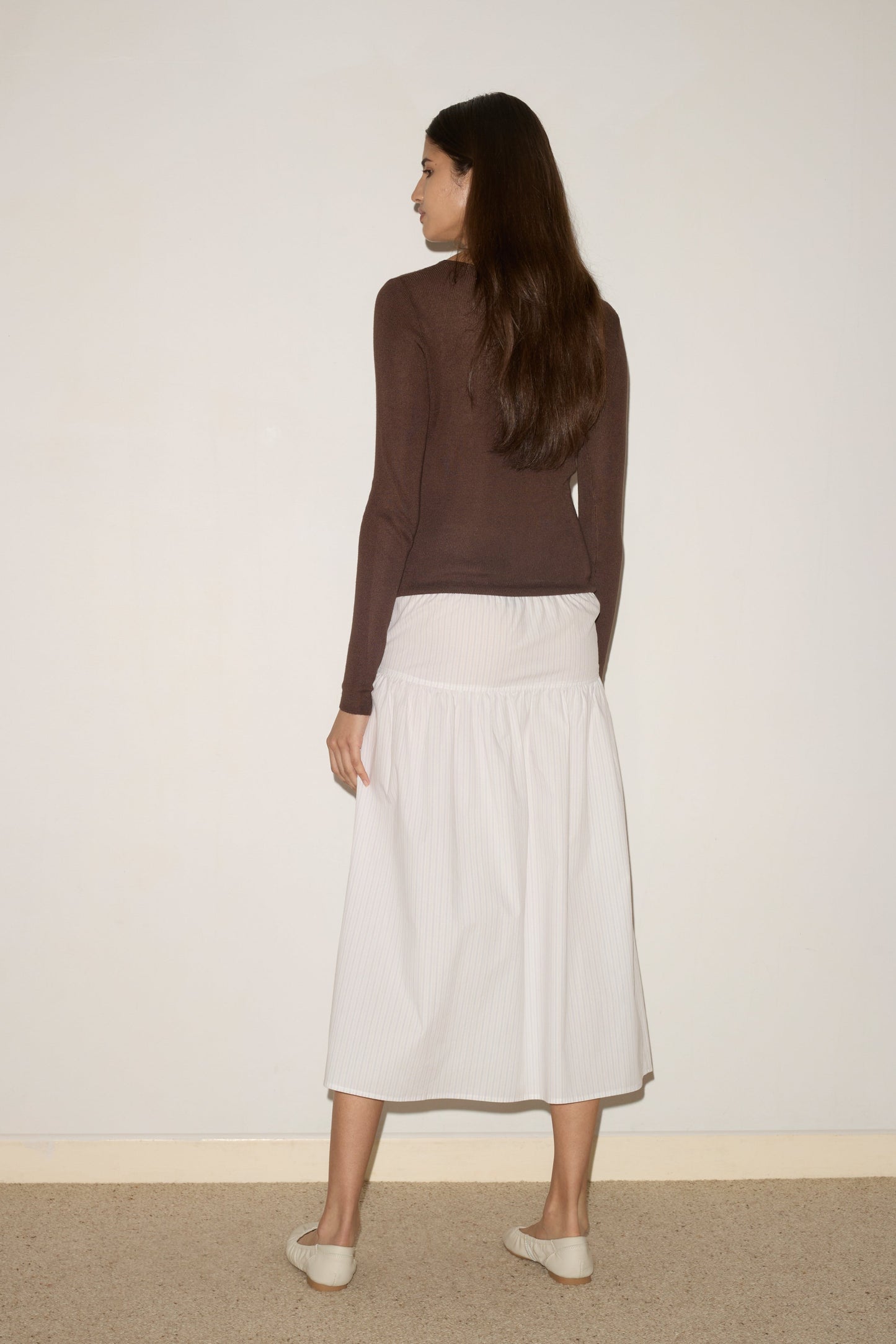 A woman with long brown hair faces a blank wall, wearing the 0 Knitted Top in dark brown, paired with a white ankle-length skirt and white flat shoes.