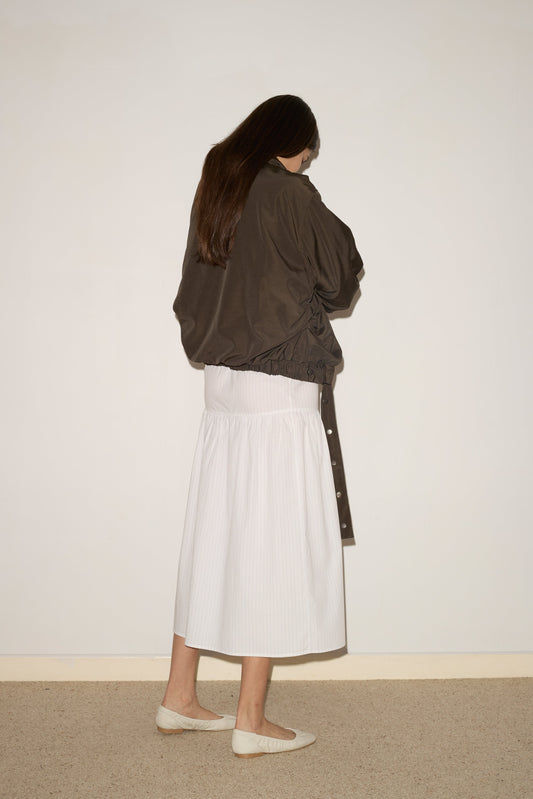 A person with long brown hair faces a wall, wearing the 0 Press Stud Jacket - Dark Chocolate, a long white skirt, and white flat shoes on a light carpeted floor against a plain off-white background.